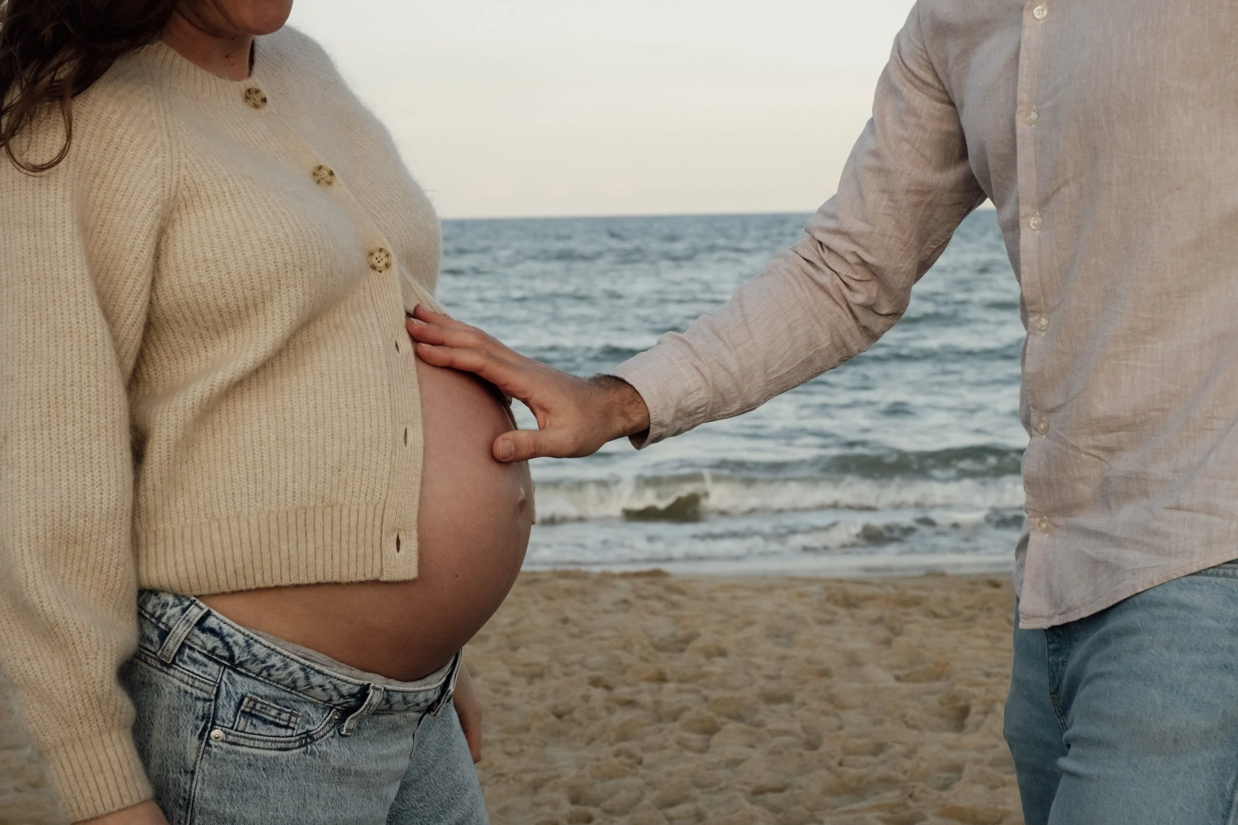 A pregnant woman with long brown hair wearing a beige cardigan with buttons and denim shorts standing on a beach with an ocean in the background. A man in a light-colored shirt with rolled-up sleeves and jeans is touching her pregnant belly.