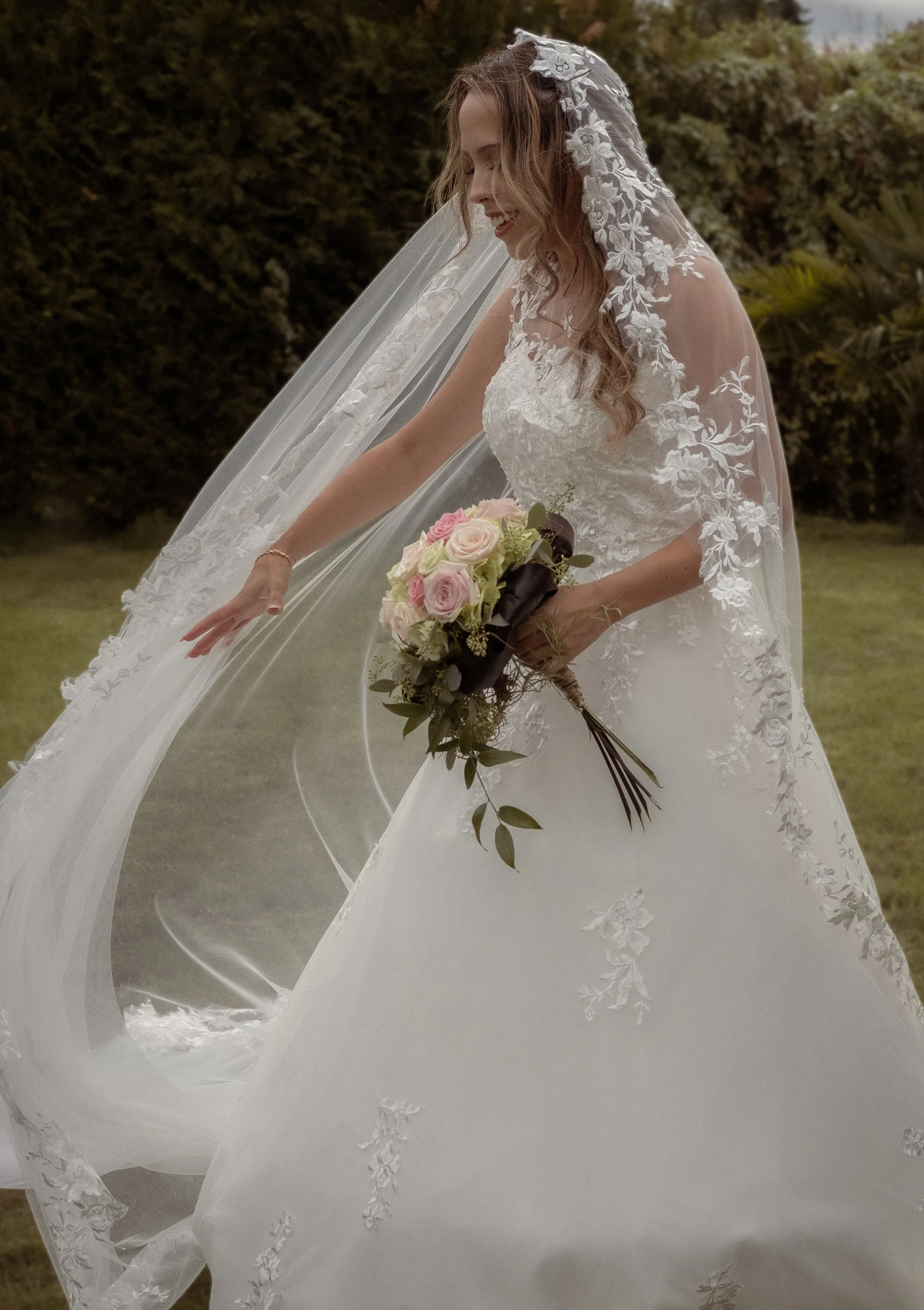 A bride wearing a white wedding gown with floral embroidery and lace, holding a bouquet of pink and white roses, with a long lace veil, outdoors in front of green bushes.