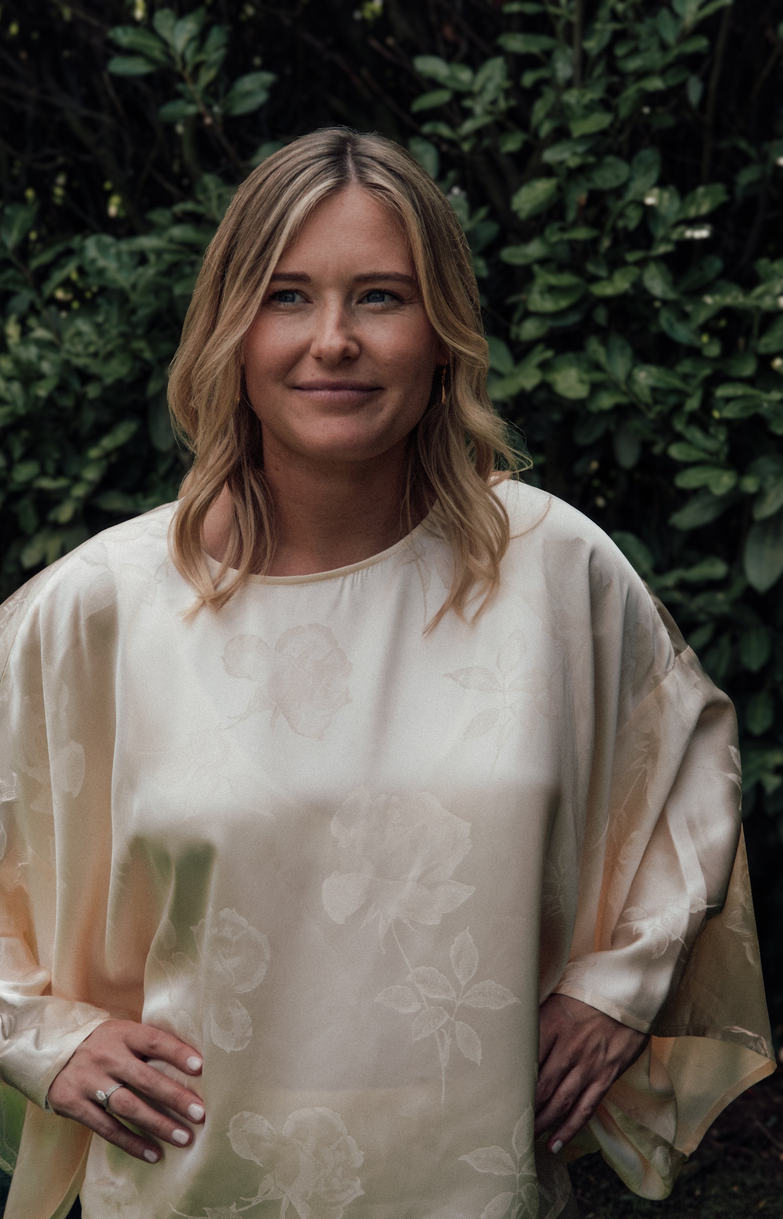Portrait of a woman with blonde hair, dressed in a cream-colored floral-patterned dress, standing outdoors among green foliage.