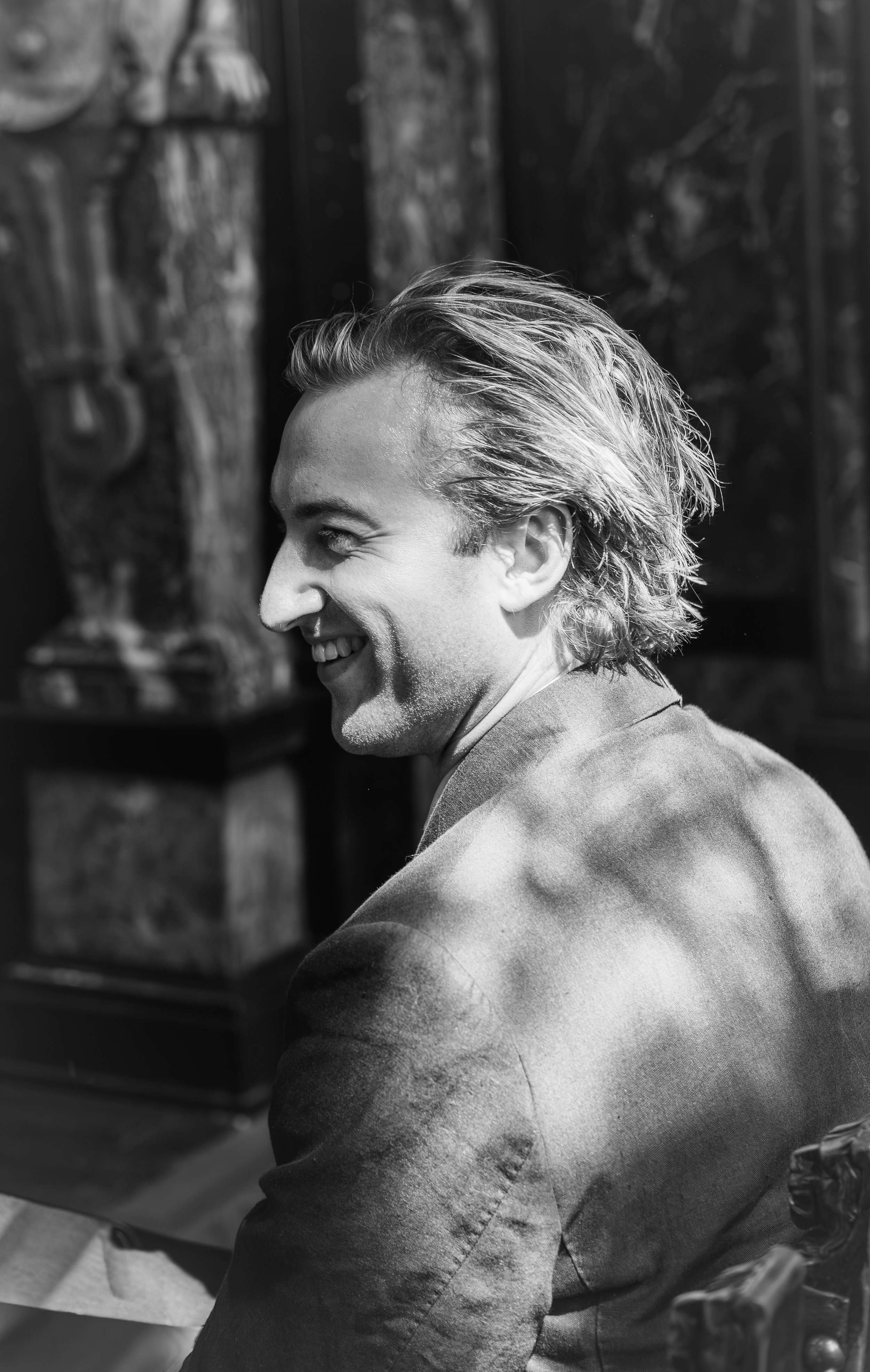 A man with light-colored hair smiling and looking to his left, wearing a light-colored blazer, in a photo taken in black and white.
