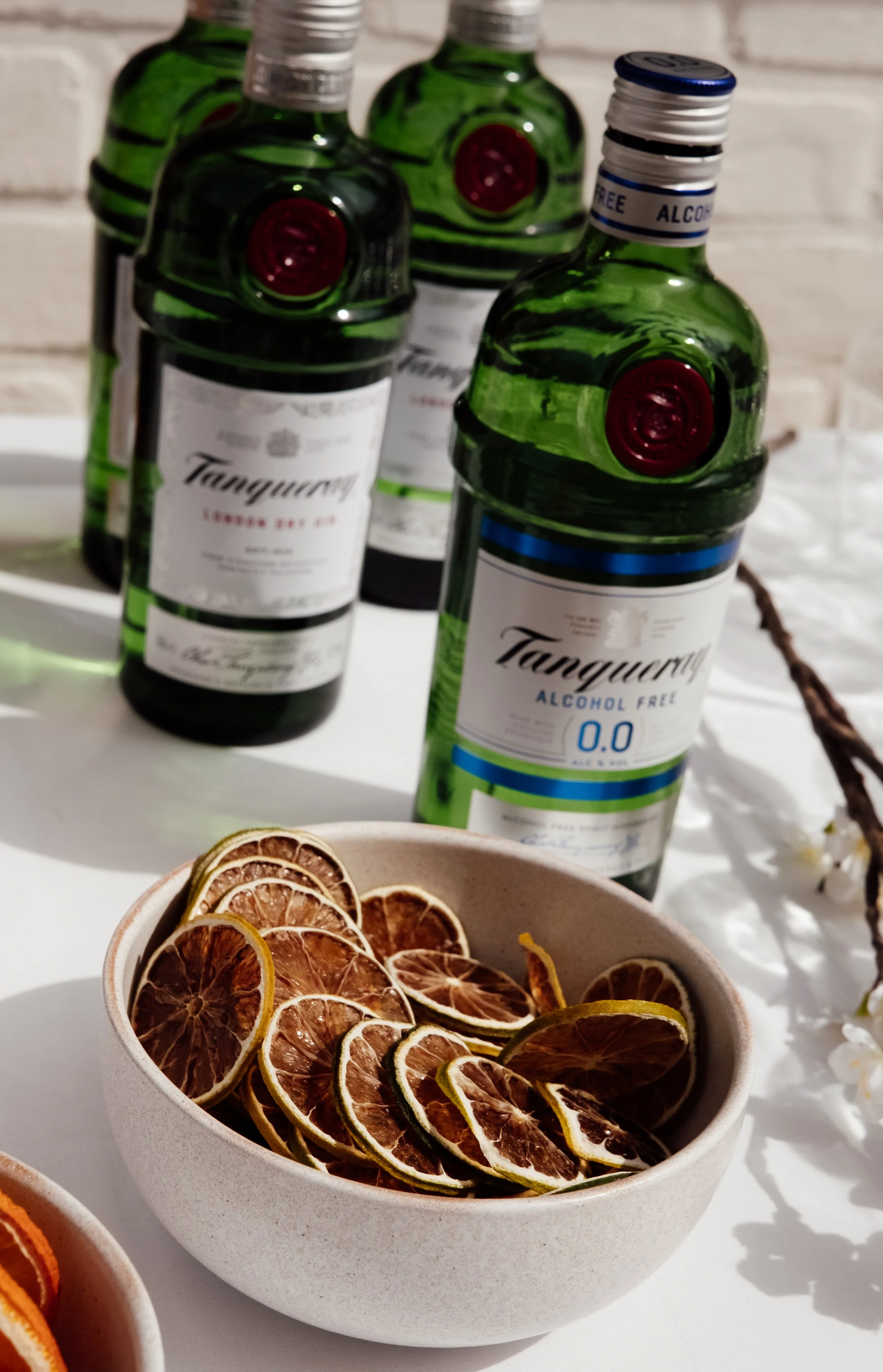 A white bowl filled with dried lemon slices in front of several bottles of alcohol, including alcohol-free tonic water, against a white brick background.