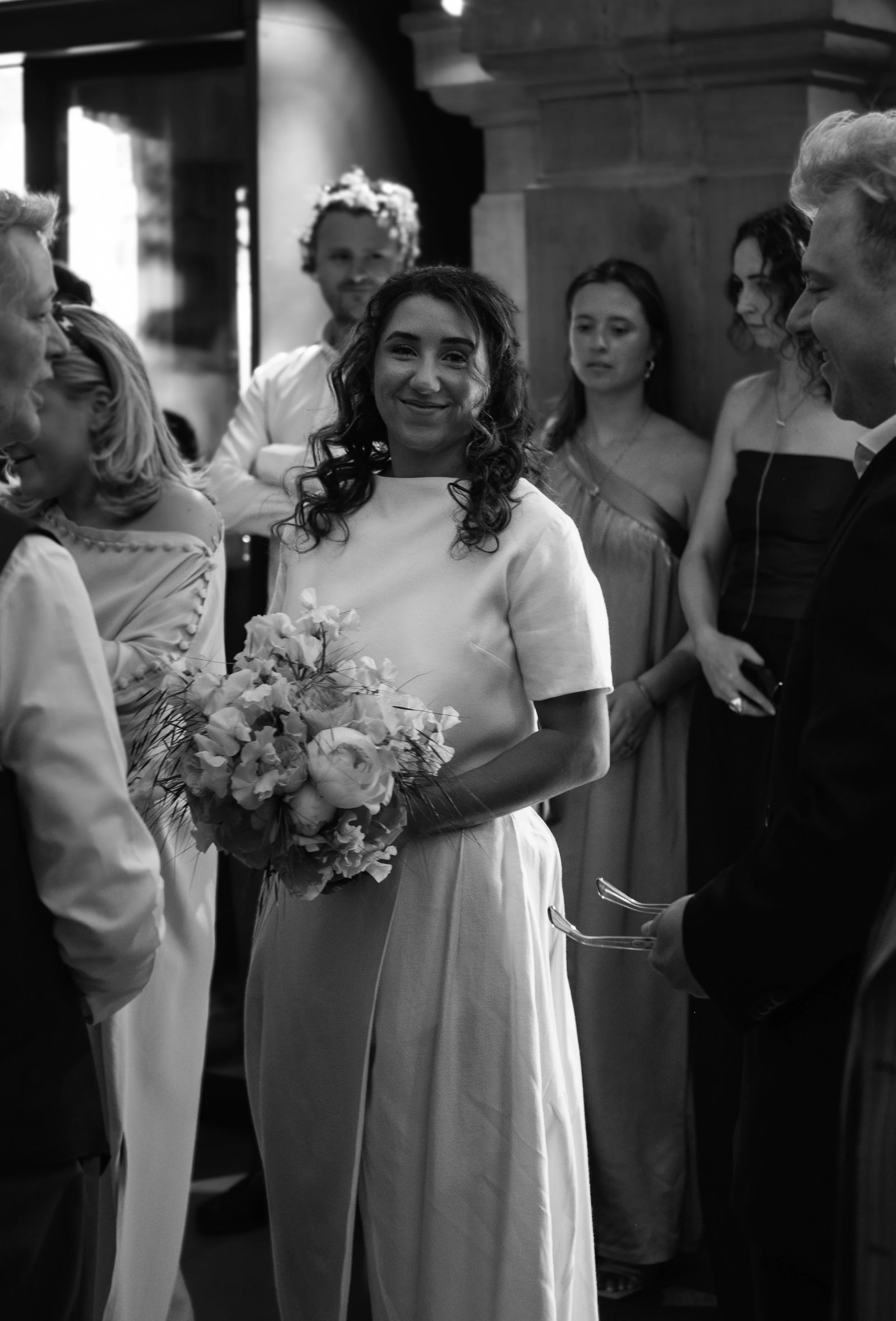 A black-and-white photo of a wedding ceremony with a smiling bride holding a bouquet of flowers, surrounded by guests in formal attire, inside a wood-paneled room.