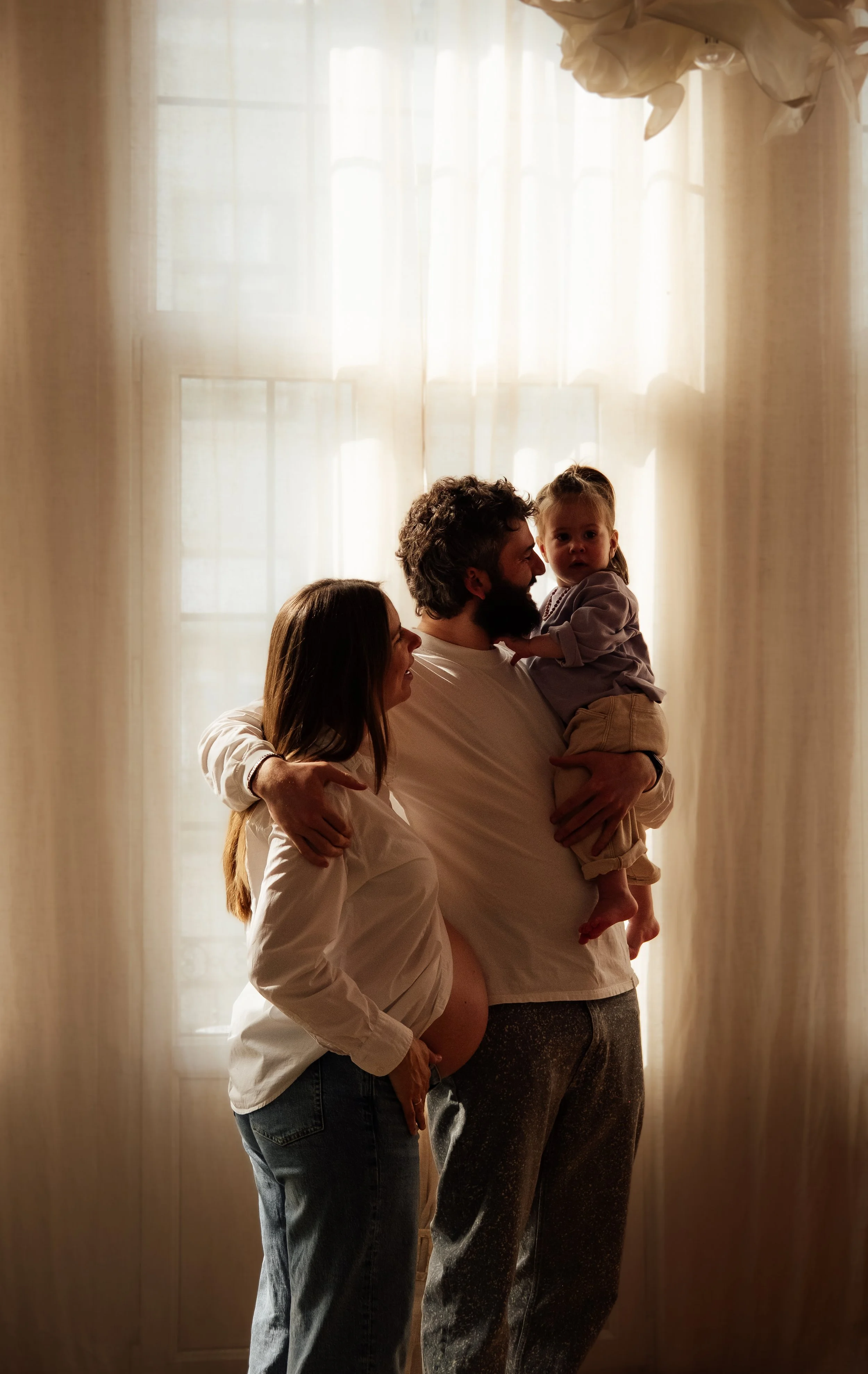 Three people, a man, a woman, and a young girl, are standing in front of a bright window. The man is holding the girl, who is looking at the camera, while the woman stands beside them, looking at the man and girl. The scene is backlit by sunlight