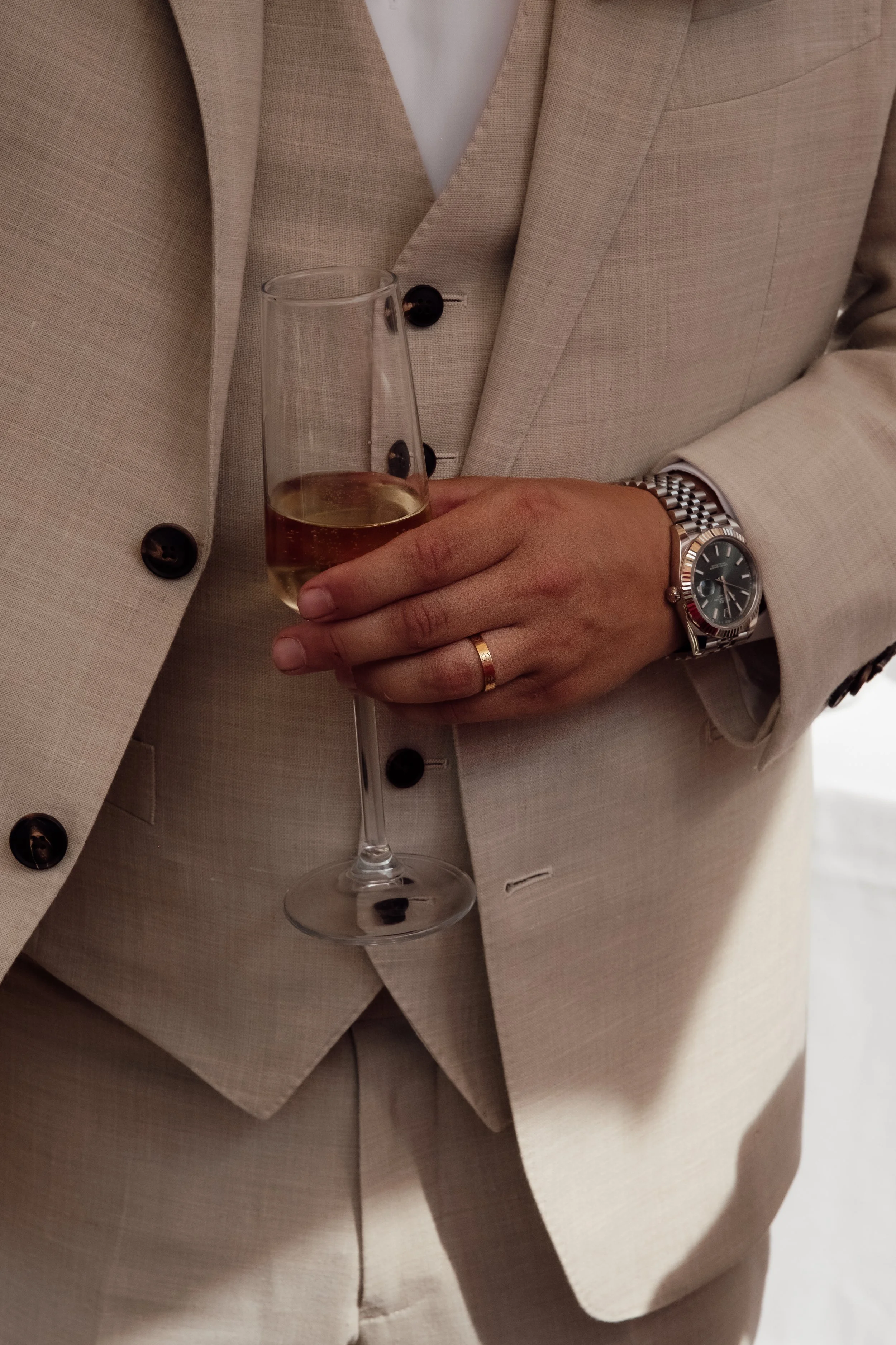 A person in a beige suit holding a champagne flute filled with champagne. The person is wearing a silver wristwatch and a wedding band.
