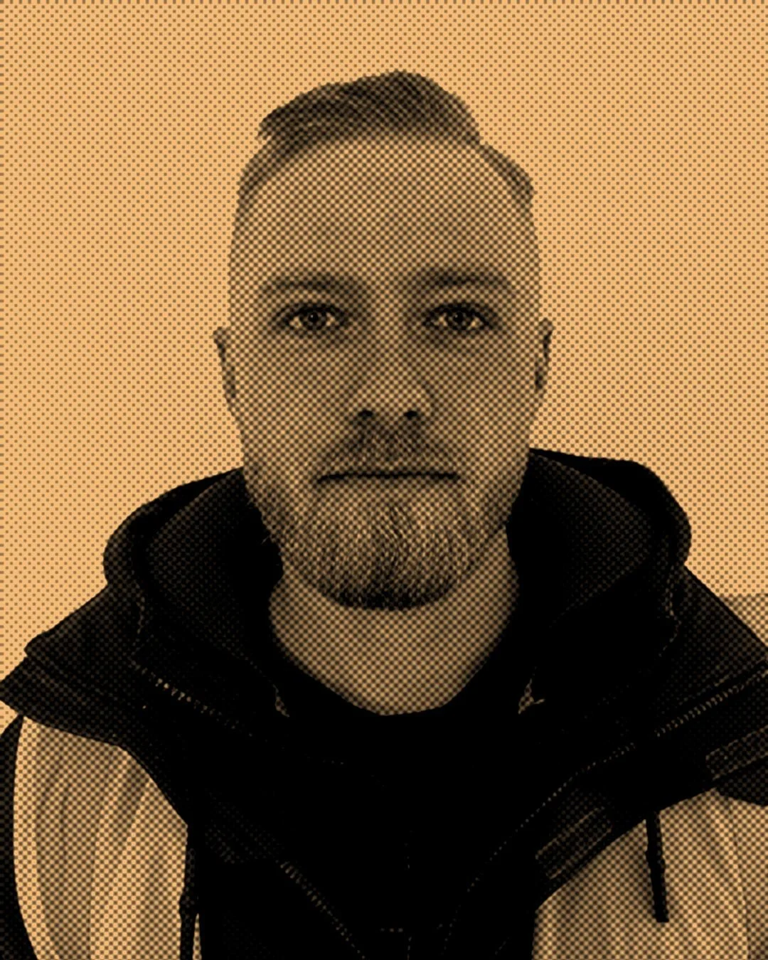 A young man with short hair and a beard, wearing a dark jacket, looking directly at the camera against a plain background.