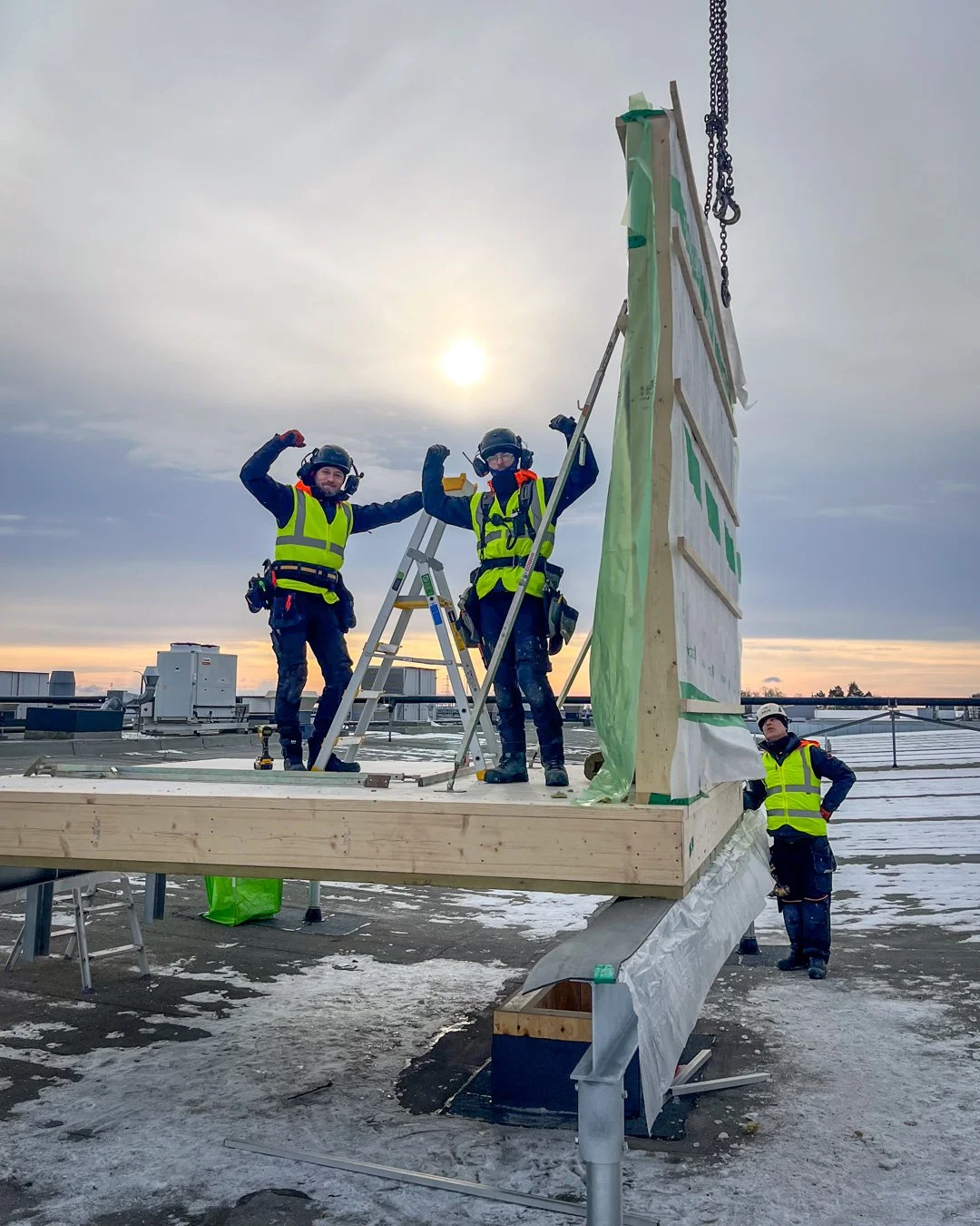 Two construction workers in safety gear and high visibility vests flex their arms on a rooftop construction site. They are standing on a wooden platform, with one worker standing beside a ladder and another near a large vertical wooden structure wrapped in green protective covering. The background features a cloudy sky with the sun partially visible and rooftop units.