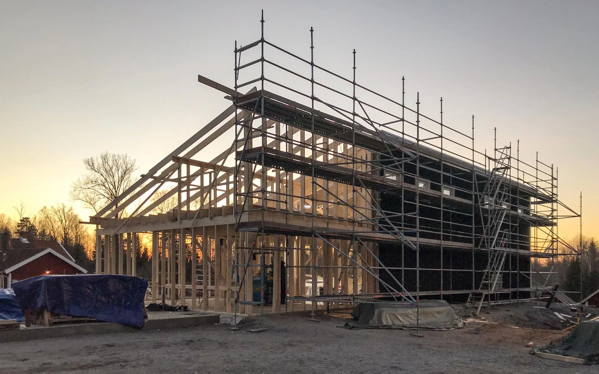 Under construction house in framings stage with scaffolding around it, during sunset night, trees in background, building materials and a tarp-covered item in foreground.