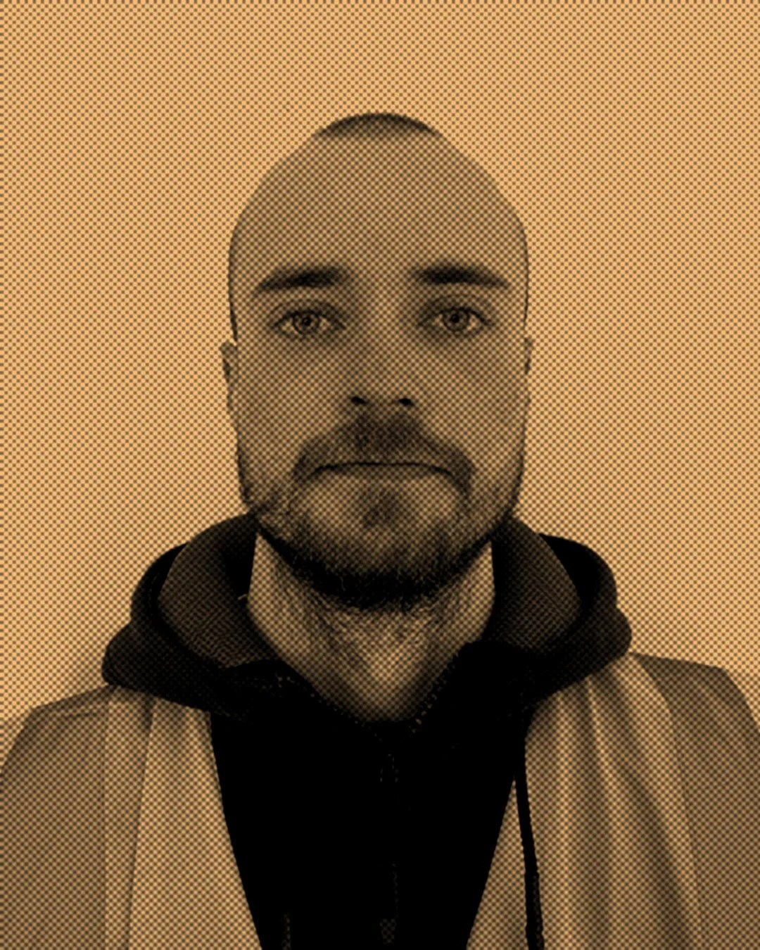 A man with a beard and bald head, wearing a hoodie and a vest, looking directly at the camera against a plain background.