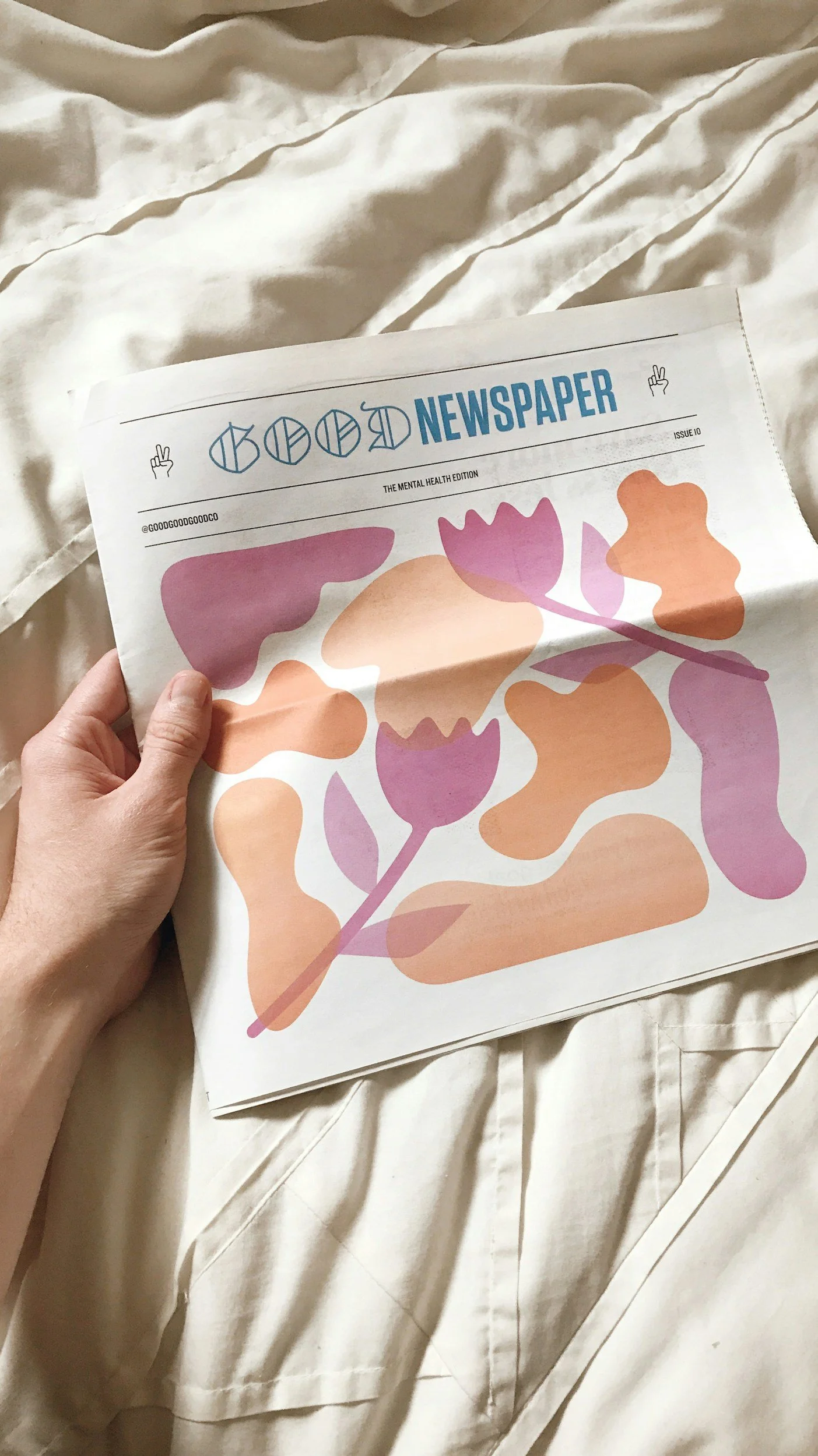 A person holding a colourful abstract themed newspaper titled on a bed with cream-colored sheets.