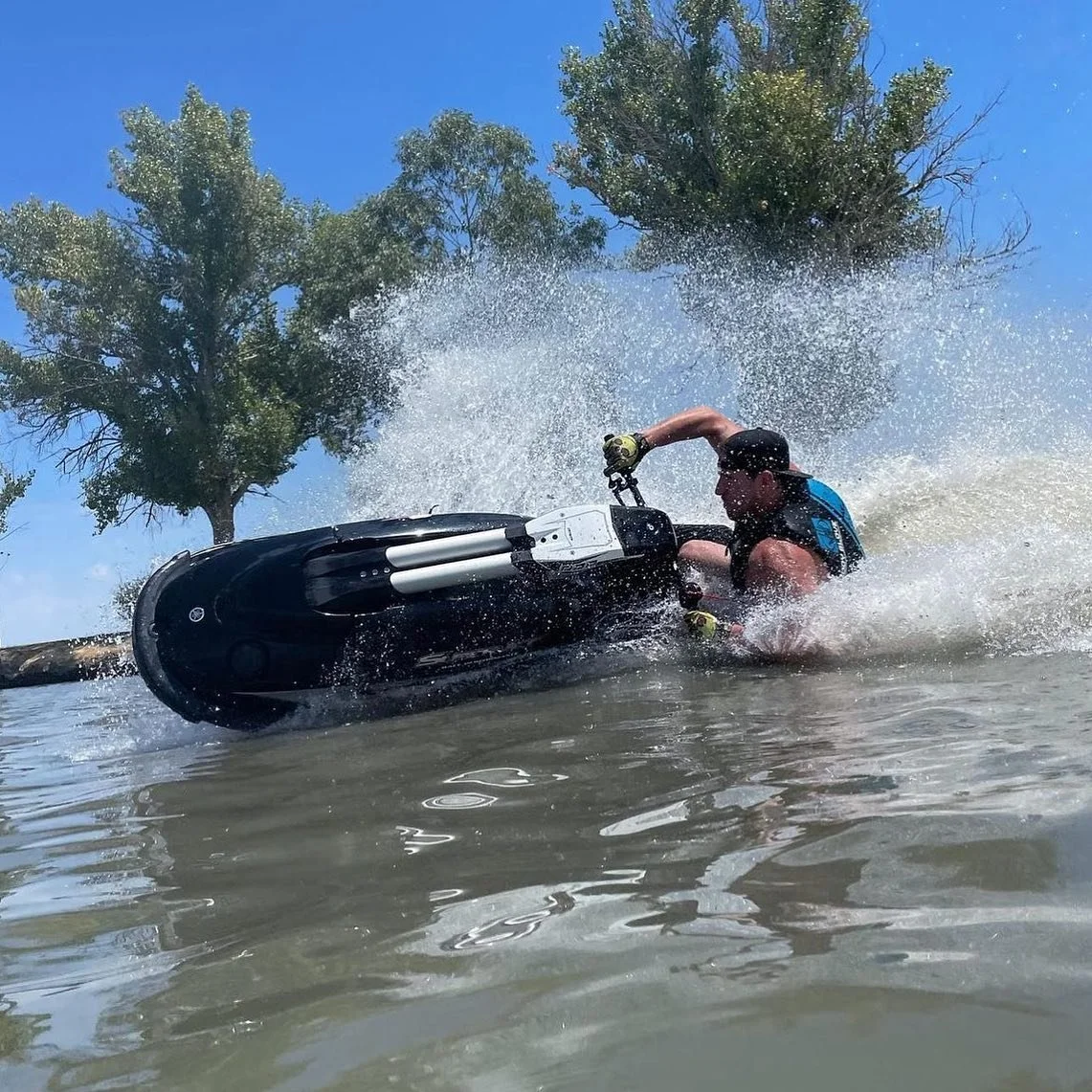 A person riding a jet ski on a body of water, kicking up spray, with trees and a blue sky in the background.