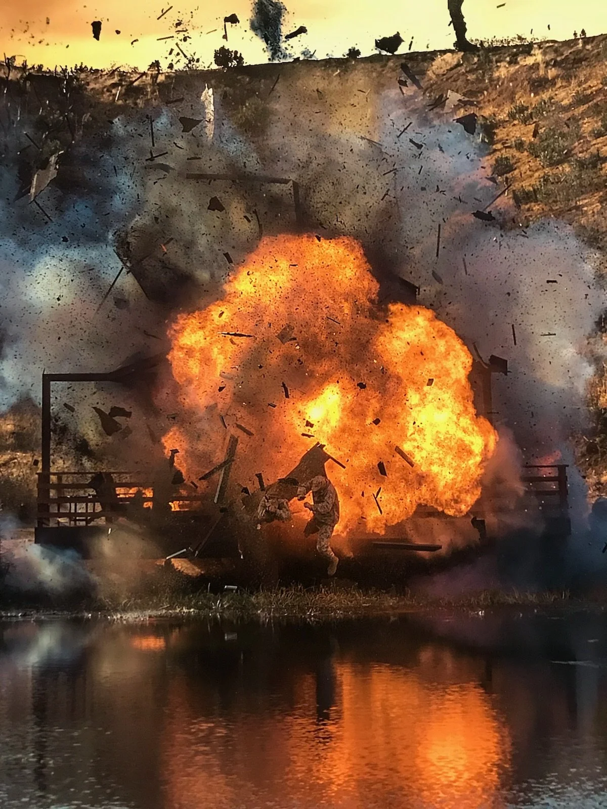 Explosion on a bridge with fire, smoke, debris, and two soldiers in a combat scene near water.