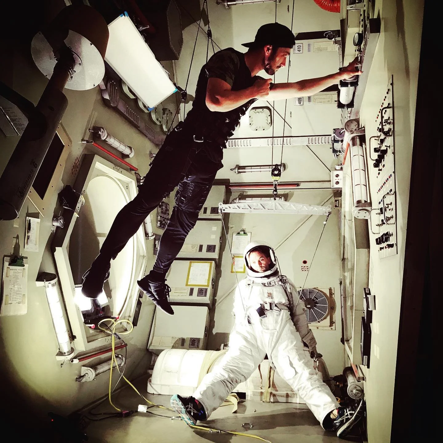 An astronaut in a white space suit floating inside the International Space Station with two crew members, one in casual attire reaching toward a control panel and another in casual clothing observing, all surrounded by equipment, cables, and scientific instruments.