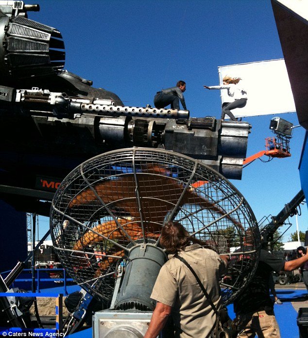 People on top of a large military tank with a mounted gun, with film crew equipment, such as a large fan and lighting, suggesting a set for a movie or photo shoot.