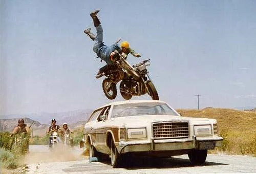 A motorcycle rider flips over a white car on a dirt road, with several motorcyclists following behind.