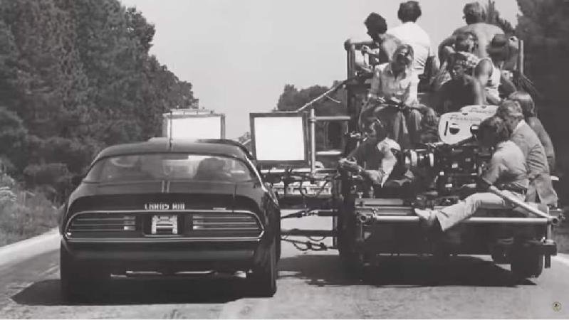 A black-and-white photo of a group of people sitting on a large filming or photography platform being pulled by a car on a road surrounded by trees.