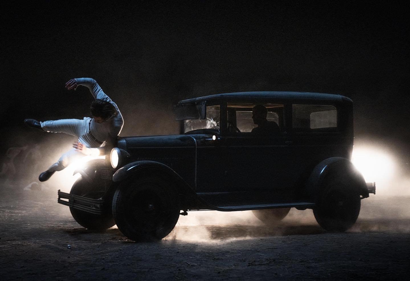 A person is falling onto the front of a vintage black car at night, with headlights on and dust or smoke in the air.