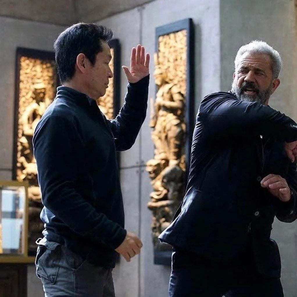 Two men in AIK jackets engaging in a friendly sparring or martial arts practice in an indoor setting, with traditional Asian-style wall art in the background.