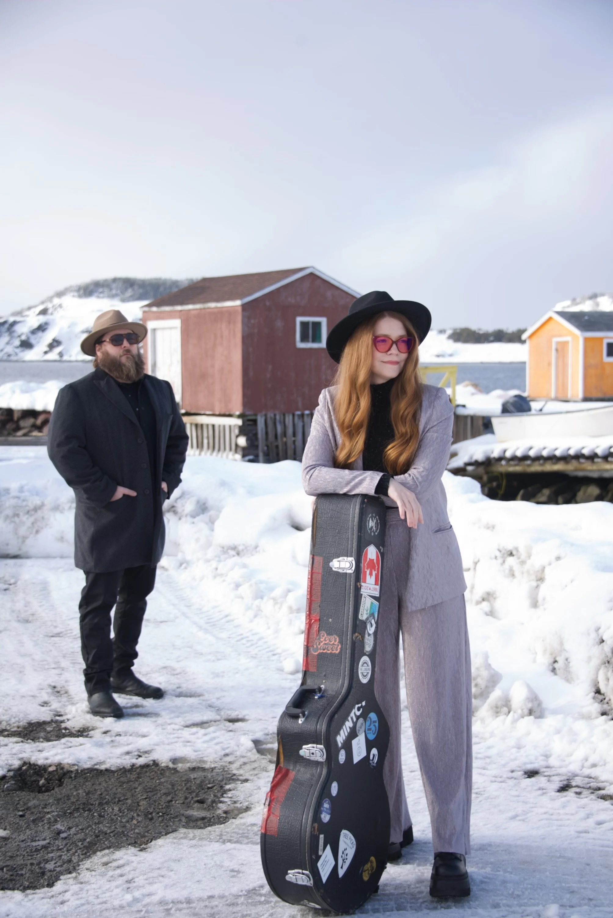 A woman with long red hair, sunglasses, and a black hat leaning on a guitar case in a snowy outdoor setting, with a man in black clothing and sunglasses standing behind her. There are small colorful houses and snow-covered landscape in the background.