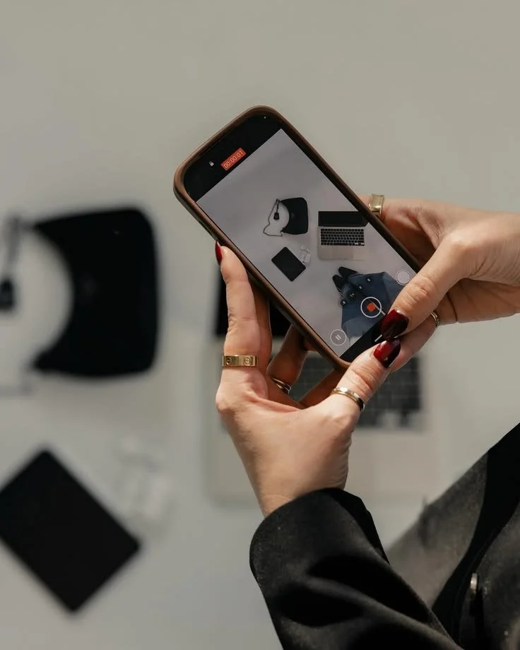 Person with painted nails holding a smartphone, recording a video of a workspace with a laptop, camera, and notebooks