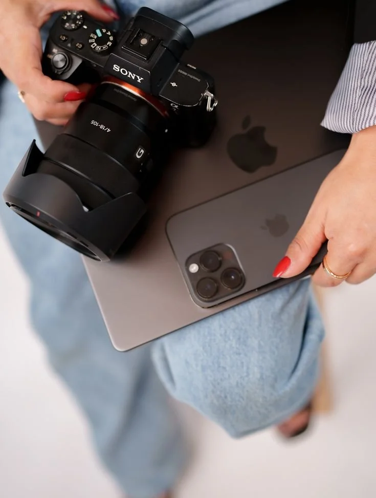 A person holding a Sony camera and an iPhone, with a MacBook in their lap, wearing blue jeans and a striped shirt.