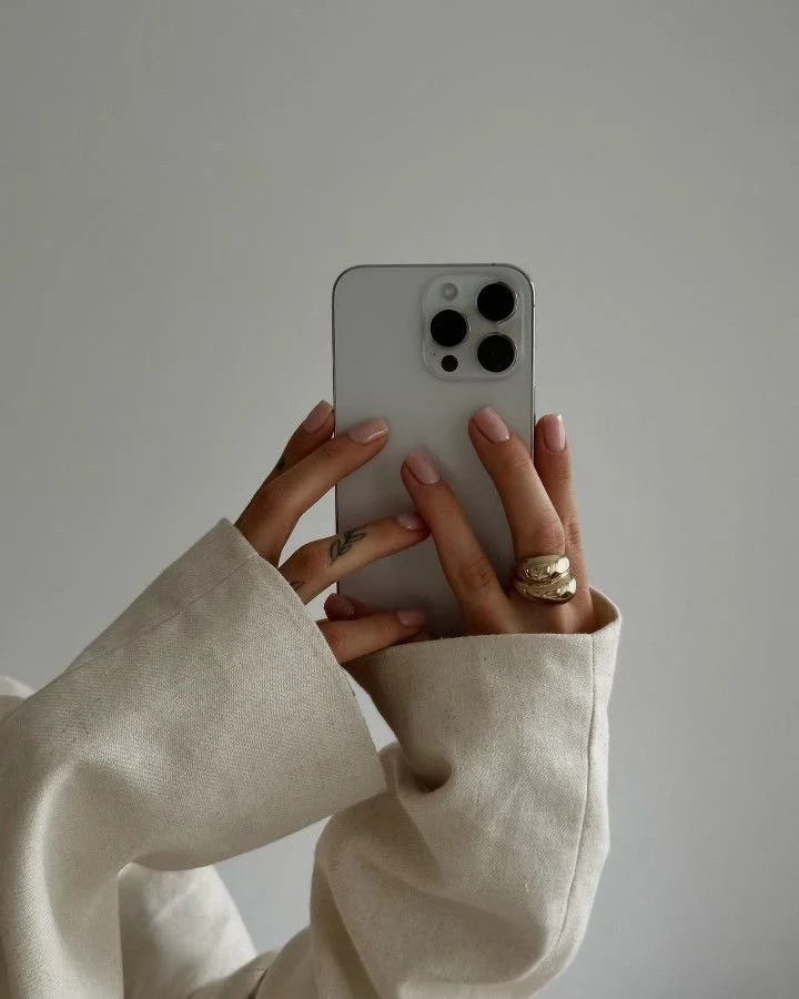 Person taking a mirror selfie with a white smartphone, wearing a beige long-sleeve shirt and rings on fingers.