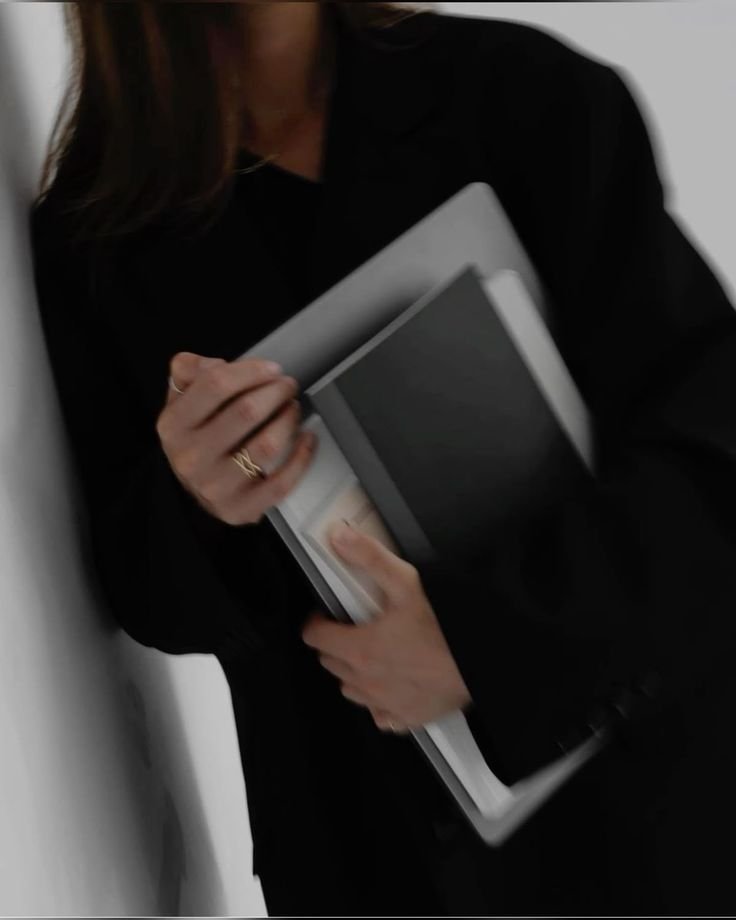 A woman in a black blazer holding a stack of notebooks or folders, partially leaning against a white wall.