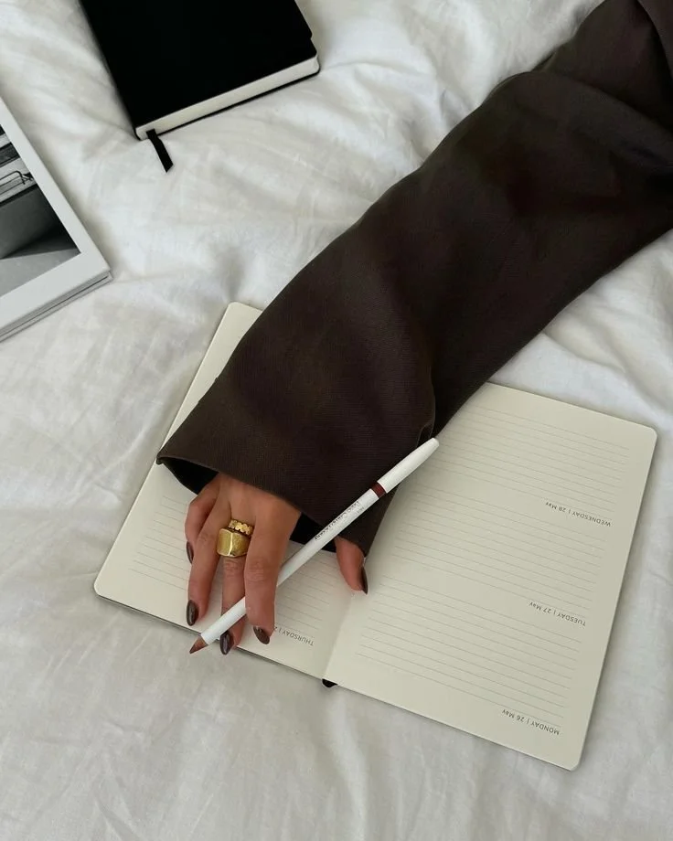 A person writing in a lined notebook with a white pen, wearing a dark long-sleeve shirt and gold rings, on a white bedspread with other notebooks nearby.