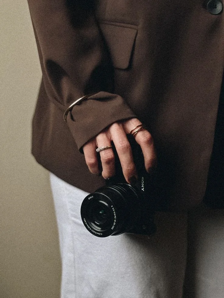 Person wearing a brown jacket, holding a Sony camera, with rings on their fingers.