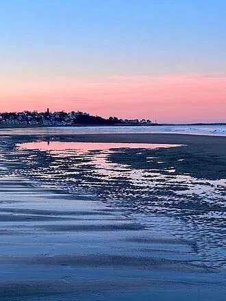 Sunset over the ocean with a sandy beach in the foreground and buildings in the distance.