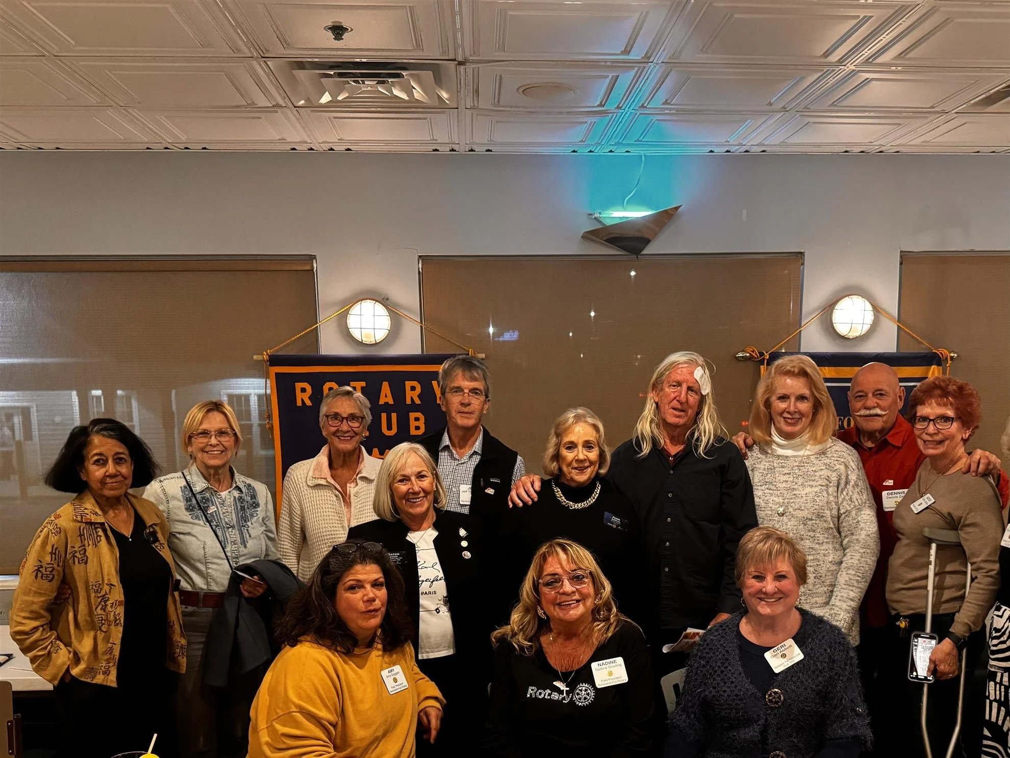 Group of people posing for a photo at a Rotary Club event, standing in front of Rotary banners, some holding name tags, in a room with beige walls and ceiling lights.