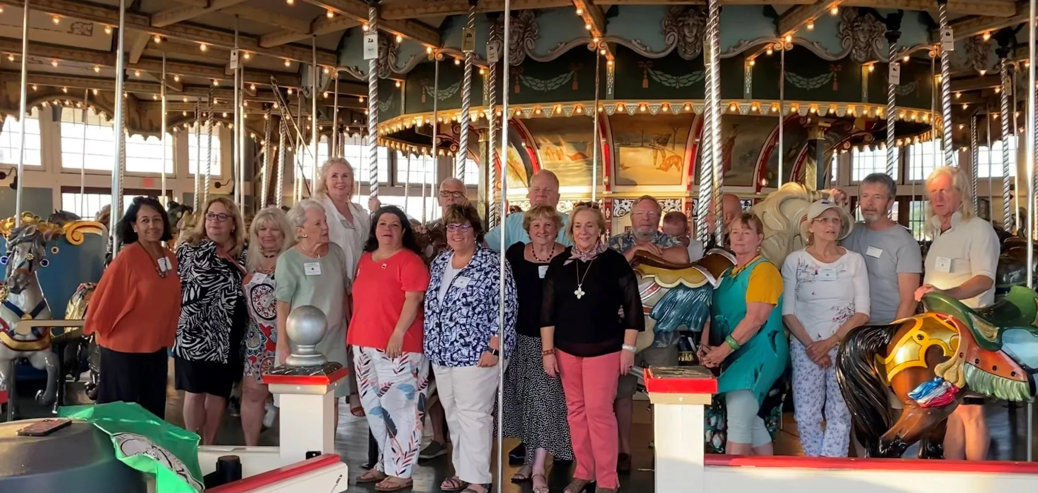 Group of people standing in front of a vintage carousel with horses inside an amusement park.