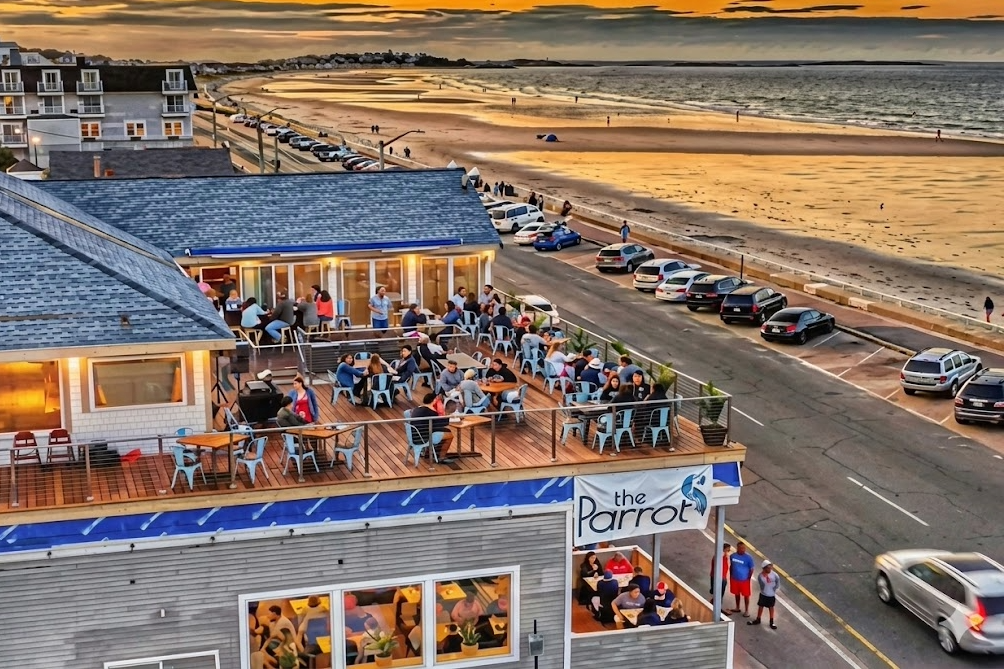 People dining outside on a wooden deck at a seaside restaurant called The Parrot, with the beach and ocean in the background during sunset.