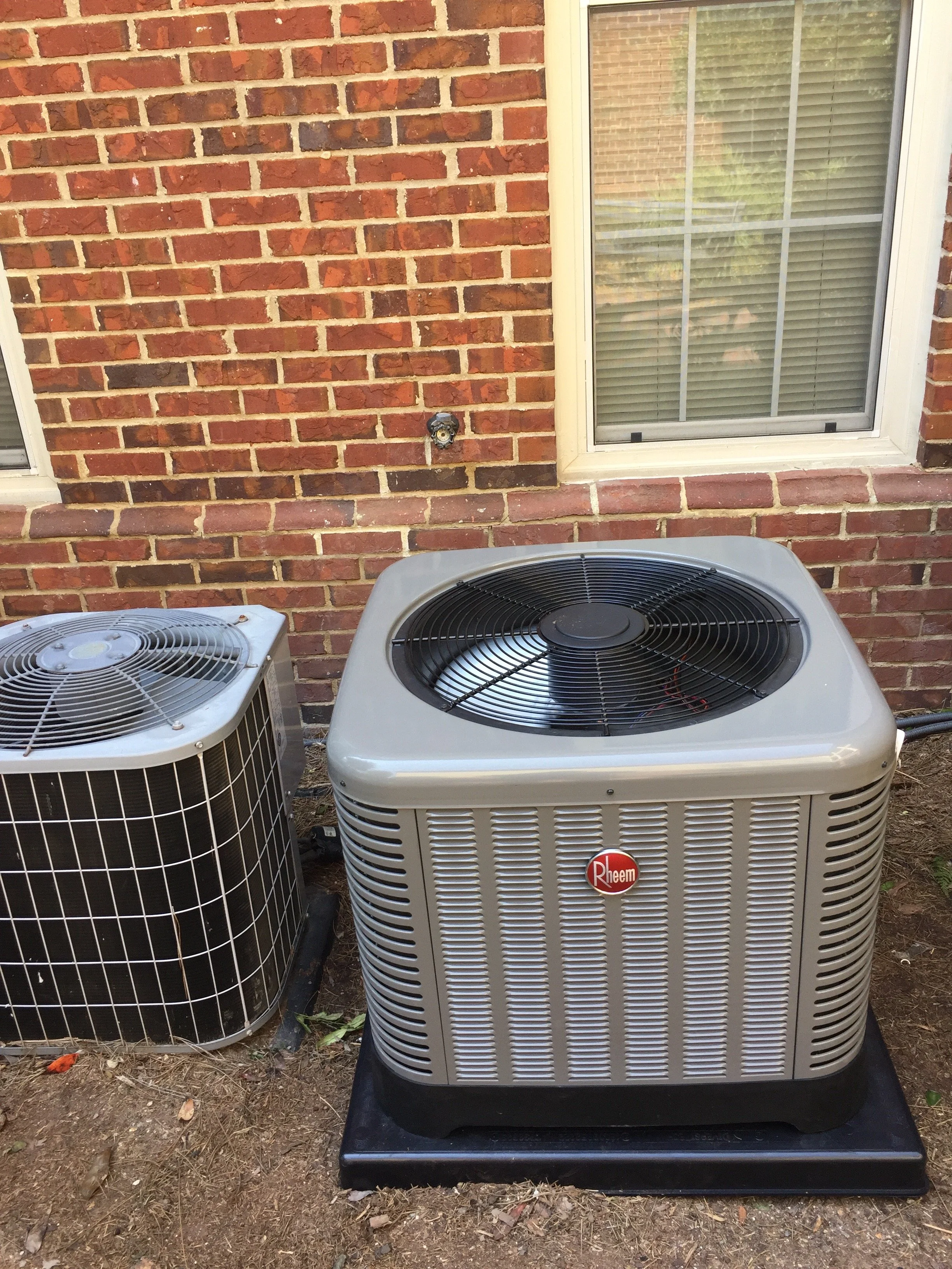Exterior view of a brick house wall with two air conditioning units on the ground outside and a window with blinds.