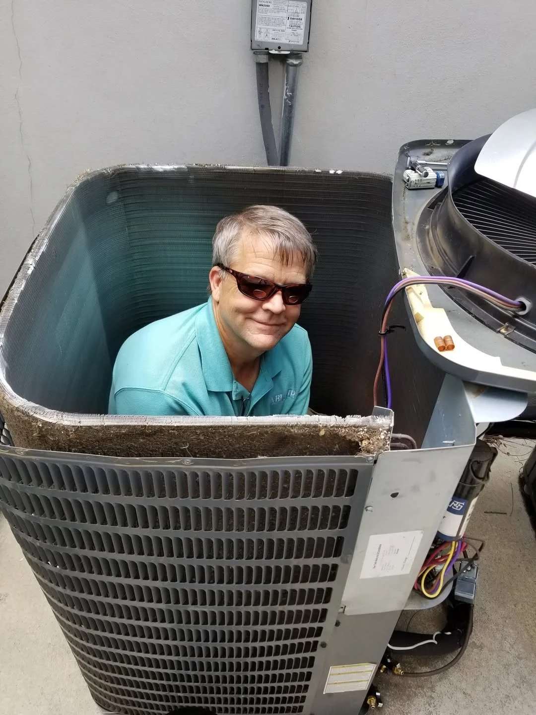 A man wearing sunglasses and a teal polo shirt is sitting inside the open, partially disassembled exterior of an air conditioning unit with electrical components and wiring visible.
