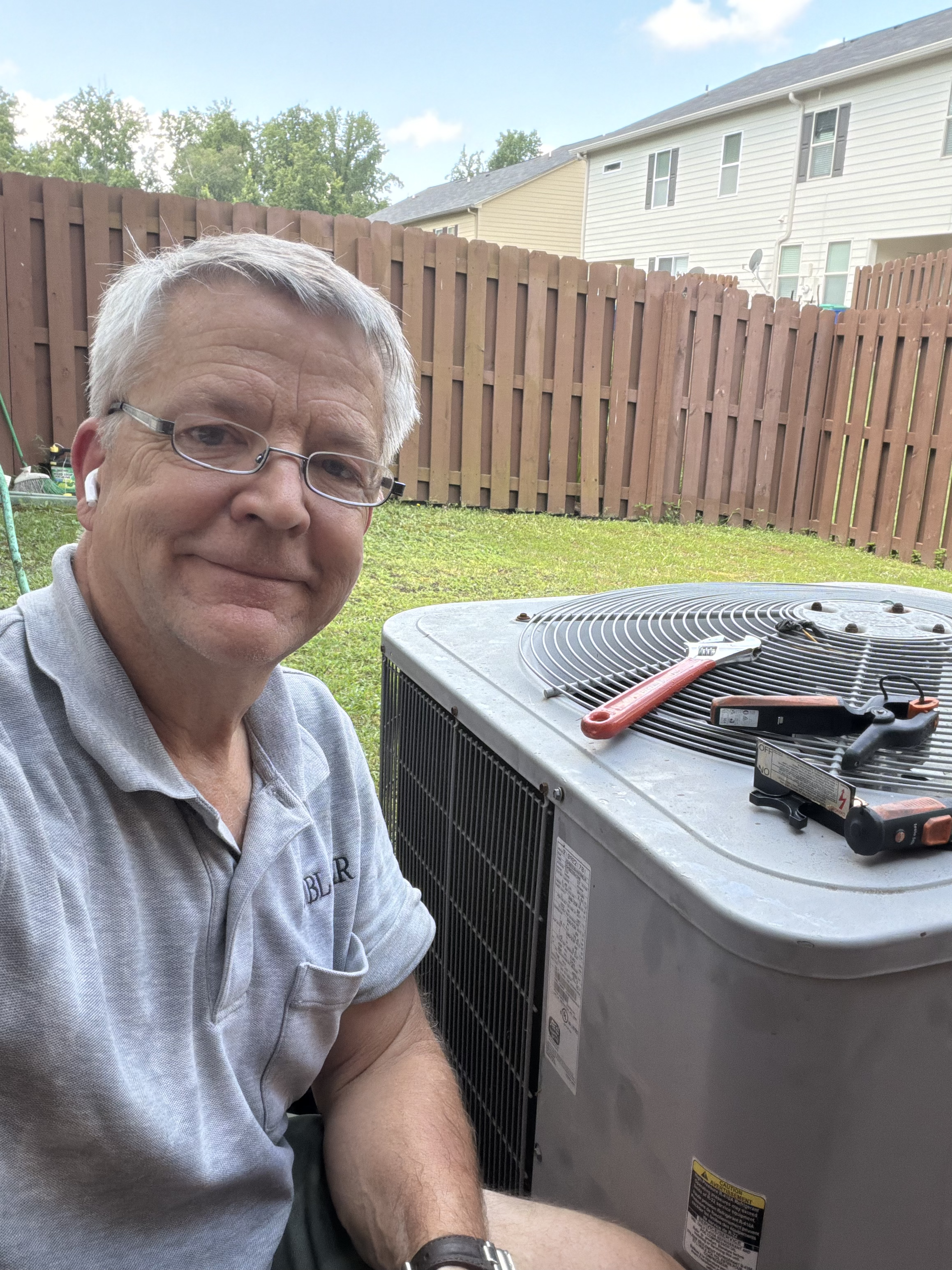 A man wearing glasses and a gray polo shirt is taking a selfie outdoors next to an air conditioning unit on a lawn, with a wooden fence, trees, and neighboring houses visible in the background.