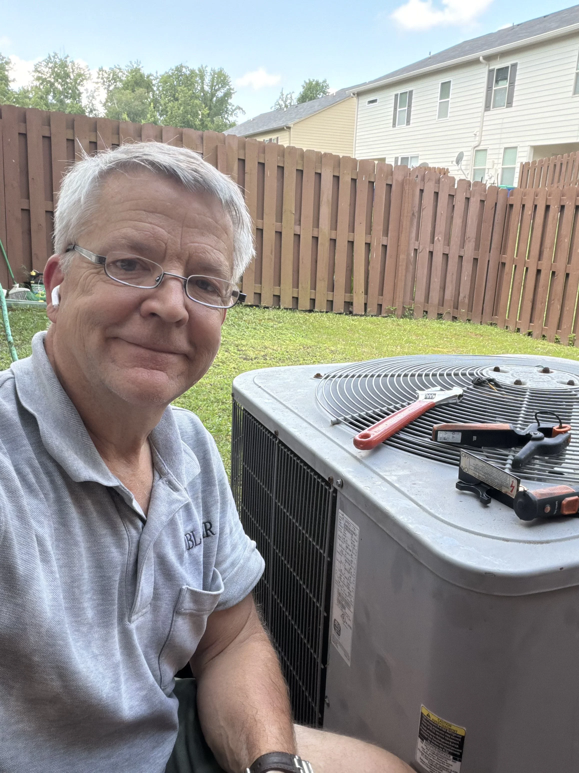 A man with gray hair, glasses, and a gray polo shirt sitting outside on a sunny day, next to a large air conditioning unit with tools on top. Fenced backyard and residential buildings in the background.