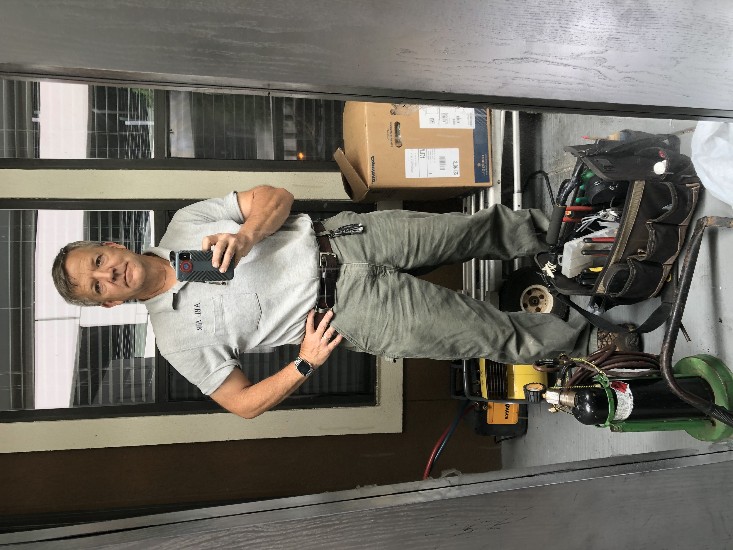 A man taking a selfie in a workshop or garage area, with tools and equipment like a toolbox, hoses, and a wheeled cart visible around him. He is wearing a light gray shirt and olive green pants, and is standing in front of a mirror.