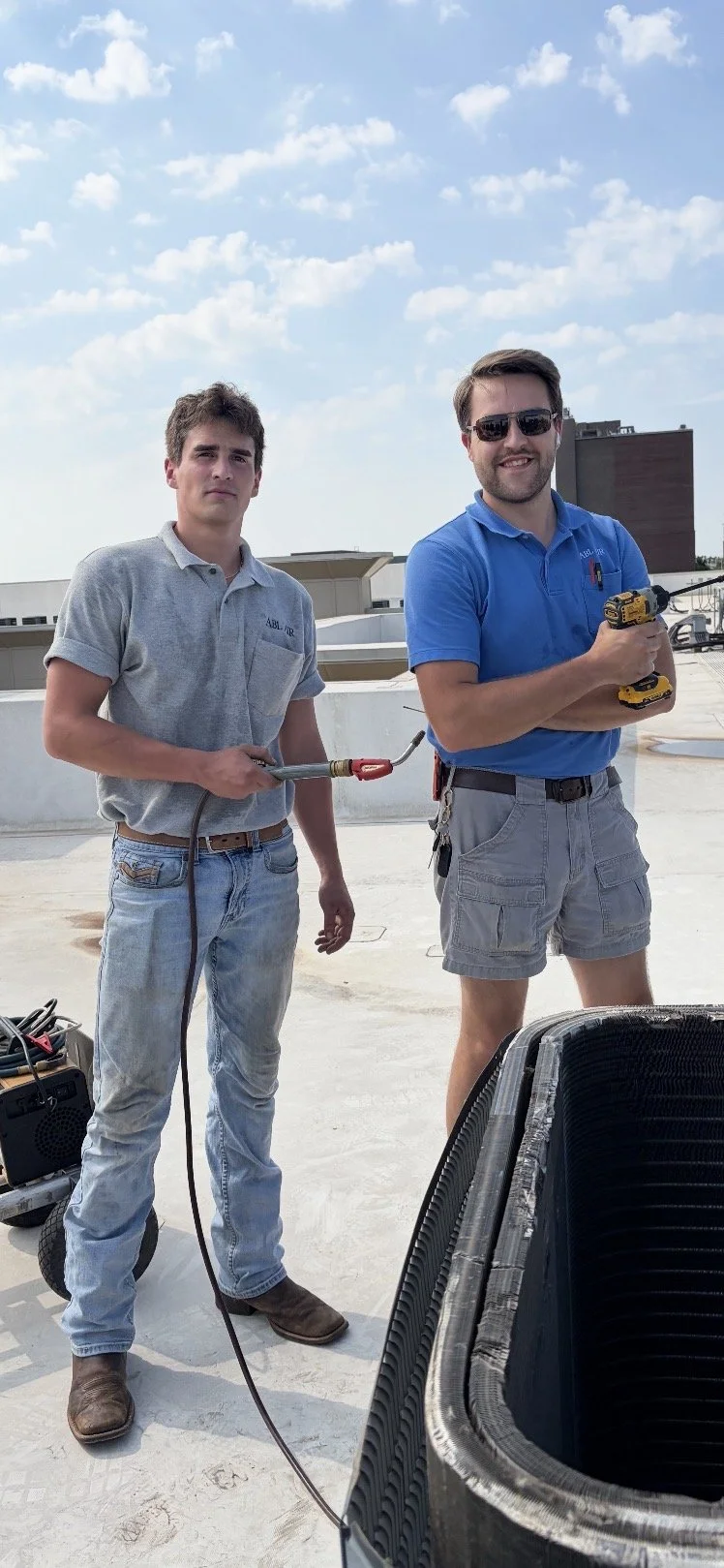 Two male workers on a rooftop under a blue sky with scattered clouds, one holding a blow torch and the other holding a cordless drill, with tools and equipment nearby.