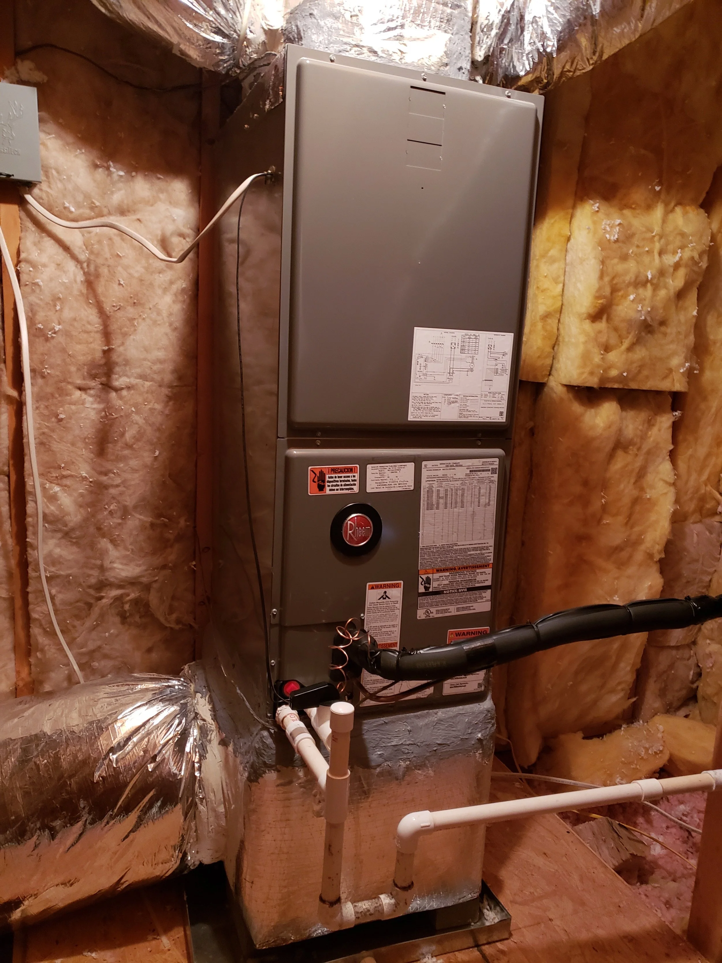 Electrical water heater installed in an unfinished basement with foam insulation and exposed pipes.