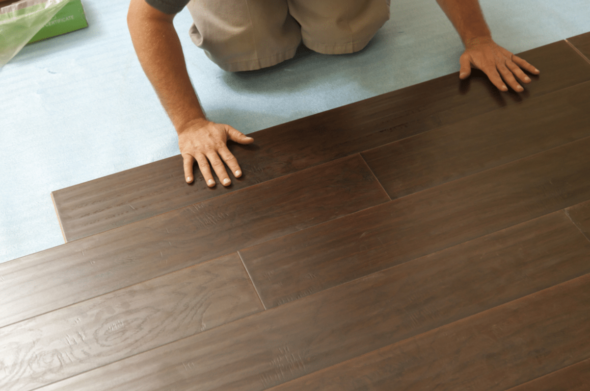 Person installing or inspecting dark brown wooden flooring on a floor, kneeling on a light-colored mat.