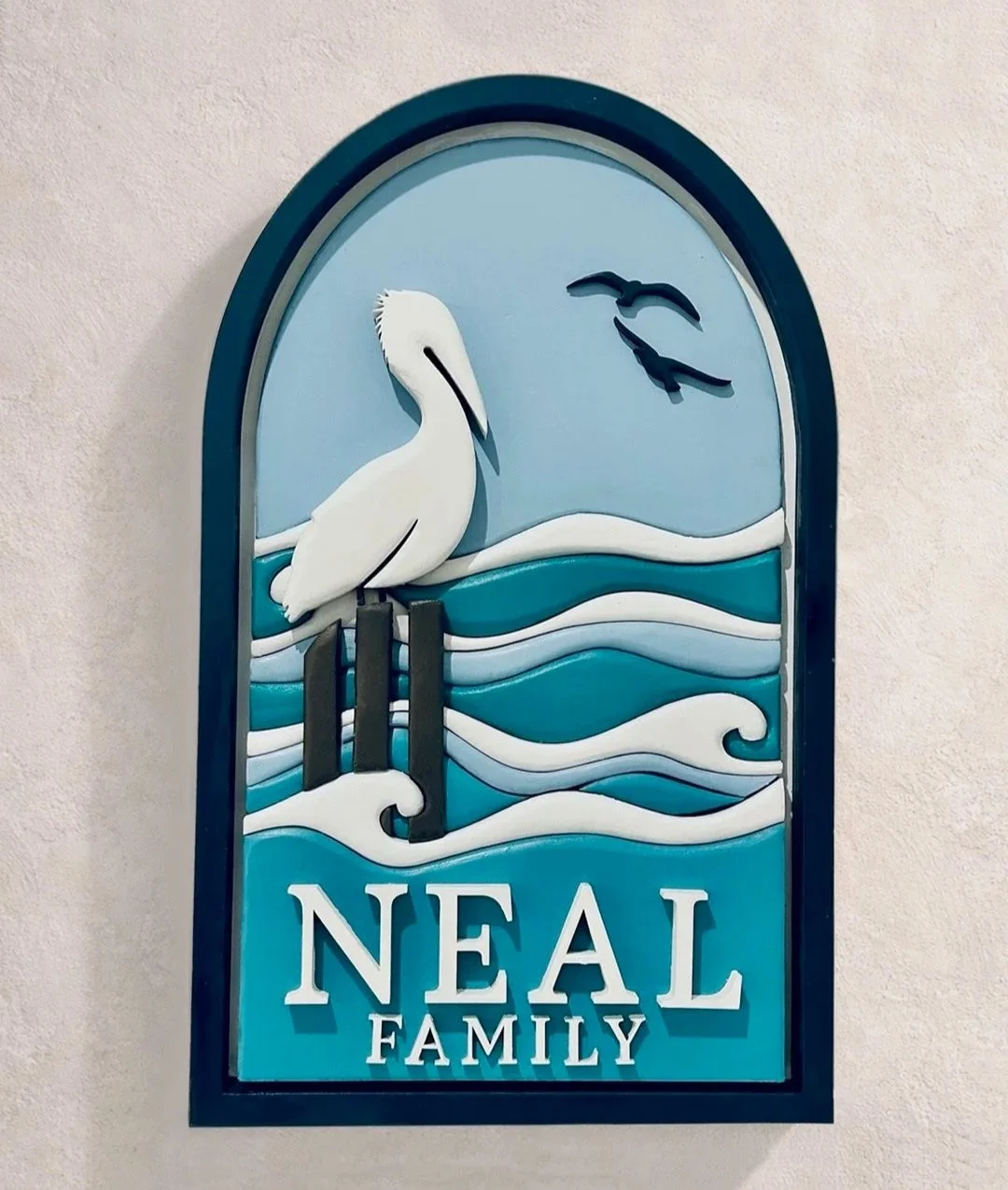 Layered family name sign reading “NEAL FAMILY” with a white heron standing over stylized blue ocean waves.