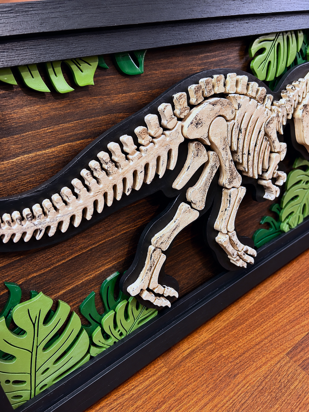 Layered wooden dinosaur skeleton wall art with tropical leaf accents and black frame.