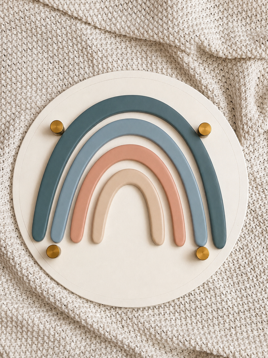 A boho-style rainbow stacker toy featuring four arched layers in muted teal, slate blue, dusty rose, and cream, mounted on a round cream acrylic board with gold brass bolts, laid flat on a textured knit blanket.