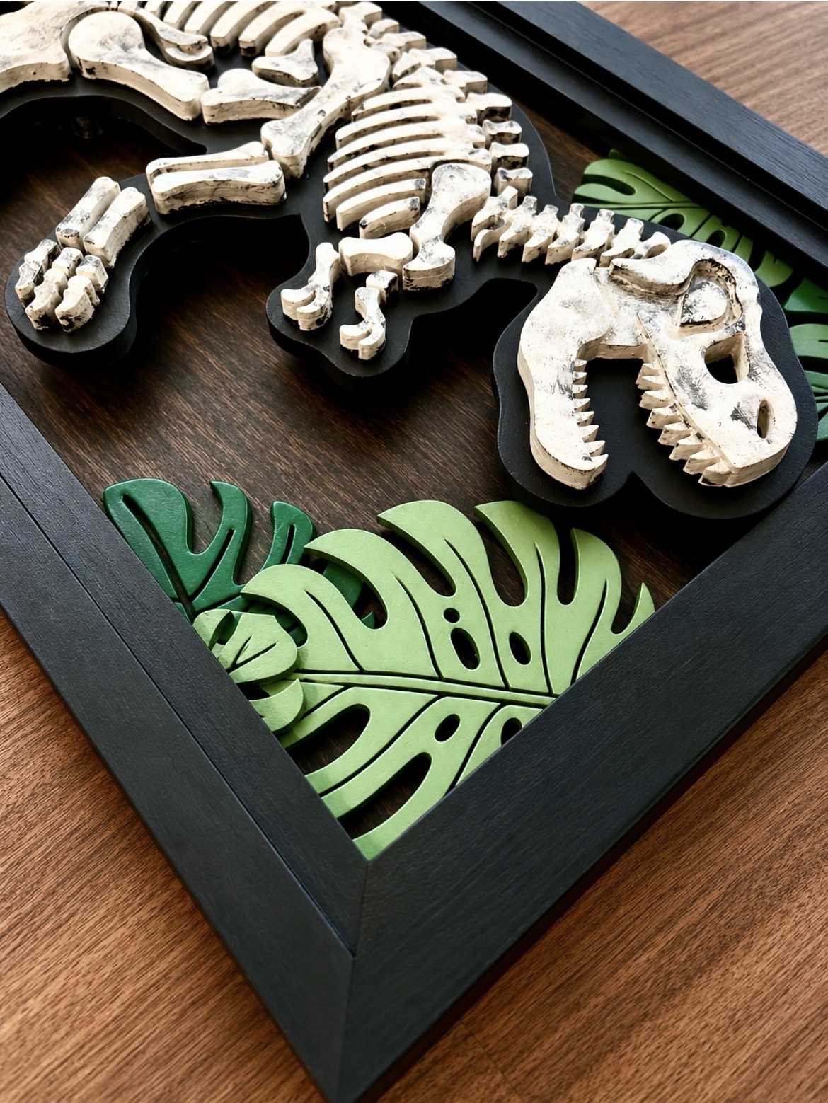 Layered wooden dinosaur skeleton wall art with tropical leaf accents and black frame.