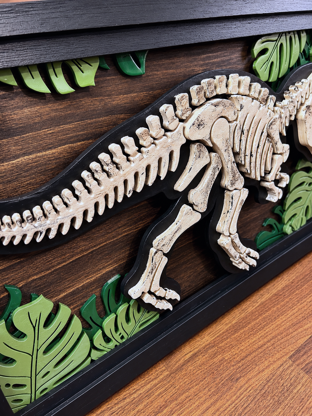 Layered wooden dinosaur skeleton wall art with tropical leaf accents and black frame.