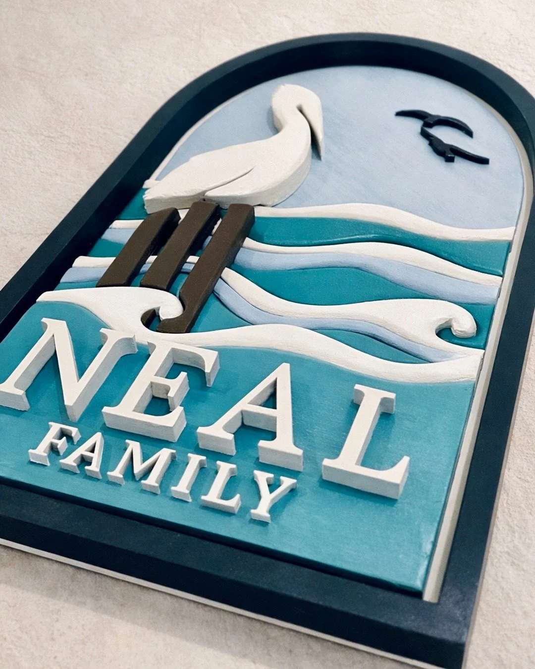 Layered family name sign reading “NEAL FAMILY” with a white heron standing over stylized blue ocean waves.