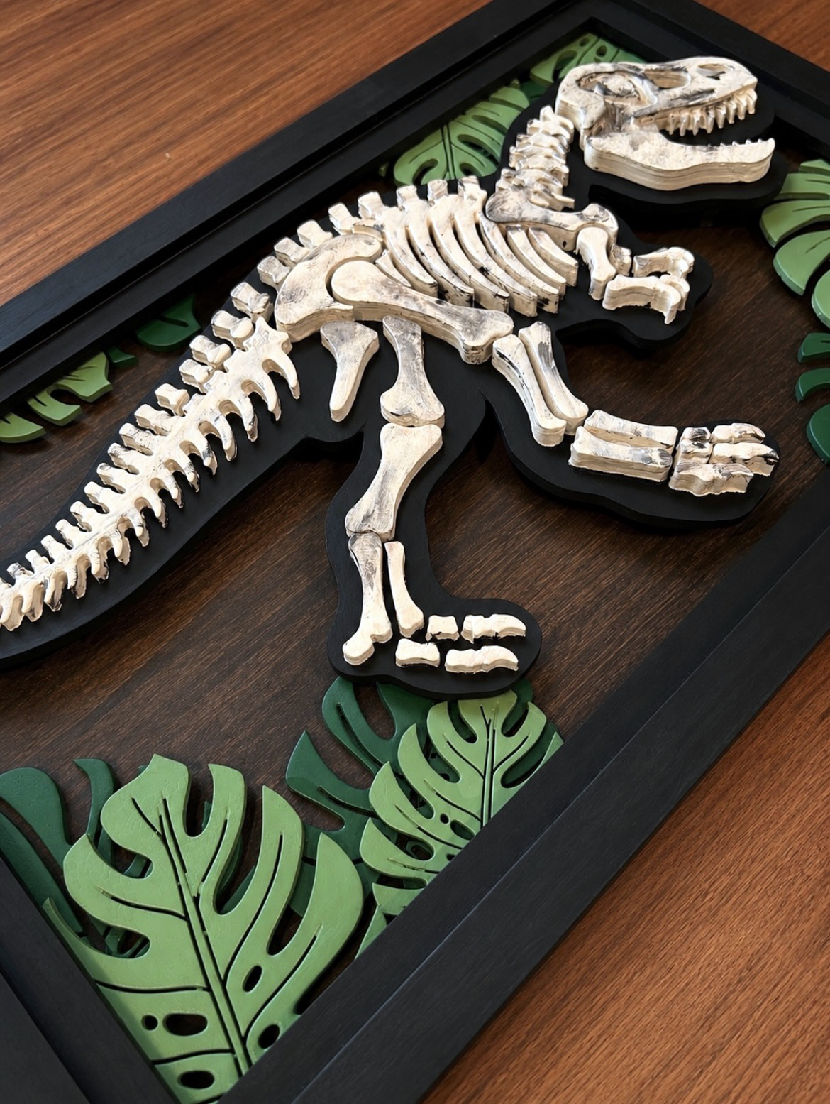 Layered wooden dinosaur skeleton wall art with tropical leaf accents and black frame.