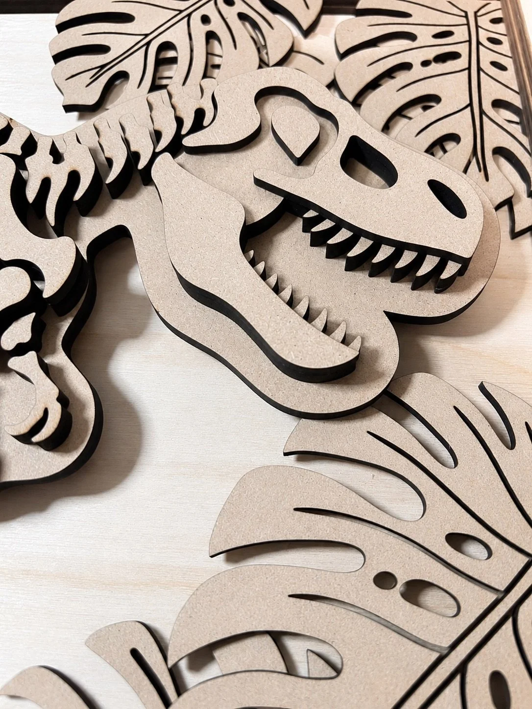 Layered wooden dinosaur skeleton wall art with tropical leaf accents.