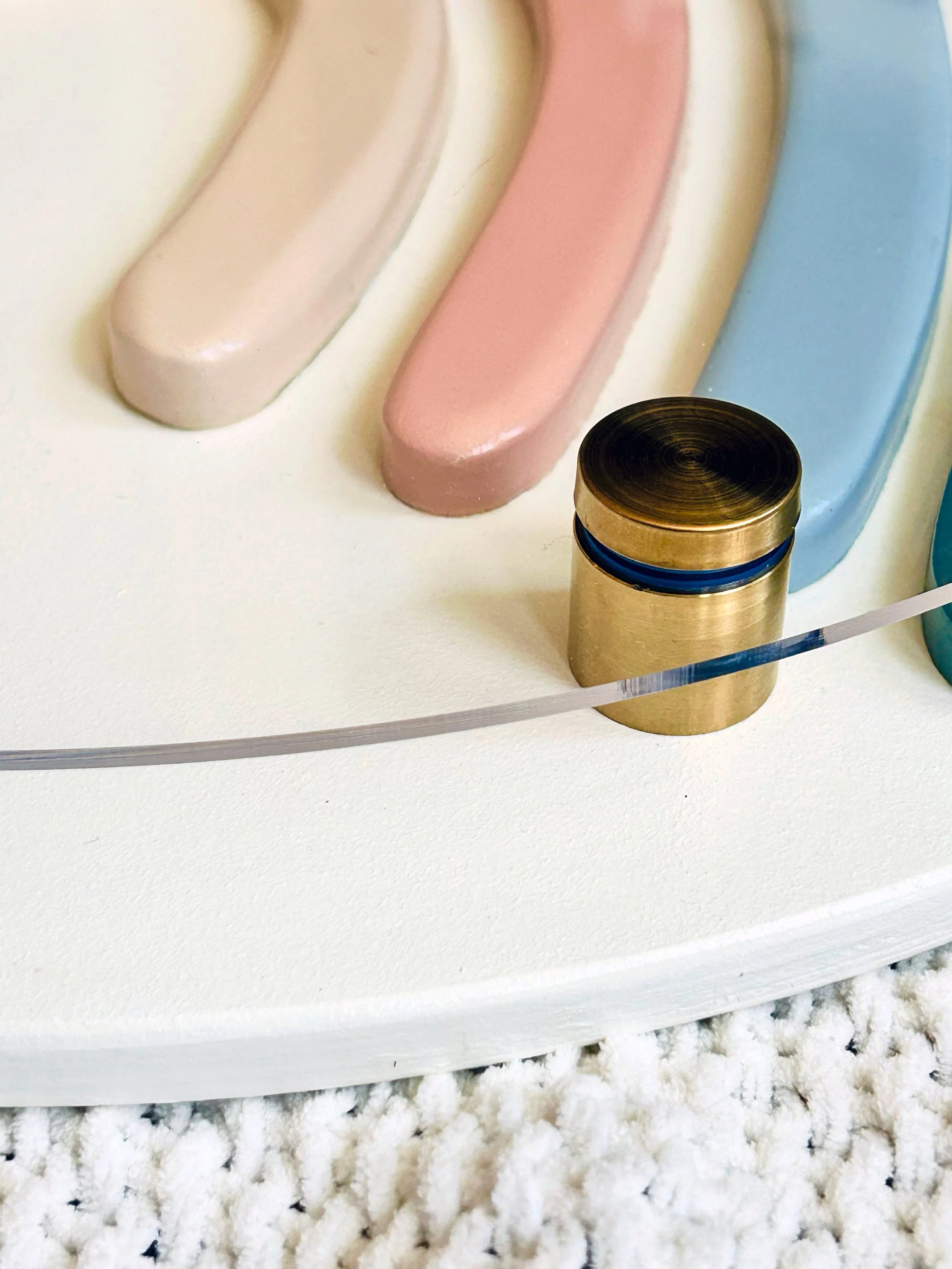 A boho-style rainbow stacker toy featuring four arched layers in muted teal, slate blue, dusty rose, and cream, mounted on a round cream acrylic board with gold brass bolts, laid flat on a textured knit blanket.