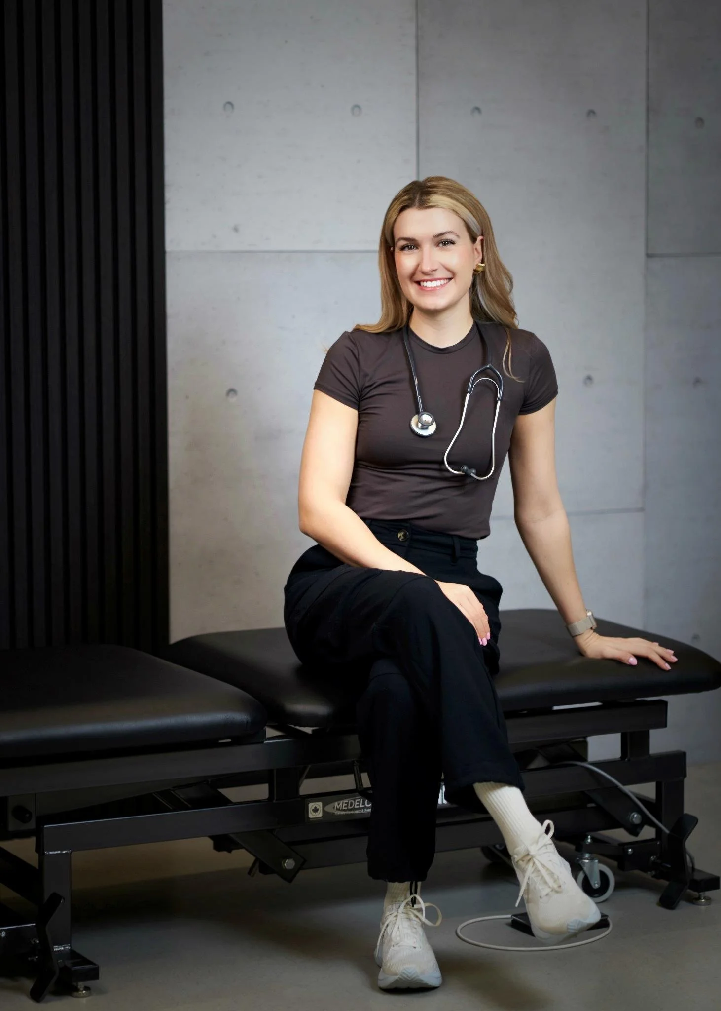 A smiling female healthcare professional sitting on an examination table in a clinical setting, wearing a stethoscope around her neck and athletic attire, with concrete and black wall backgrounds.